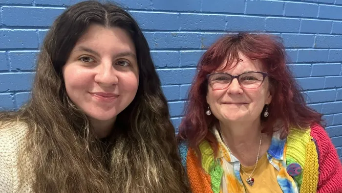 A woman with dark hair sits next to a woman with red hair in front of a blue brick wall
