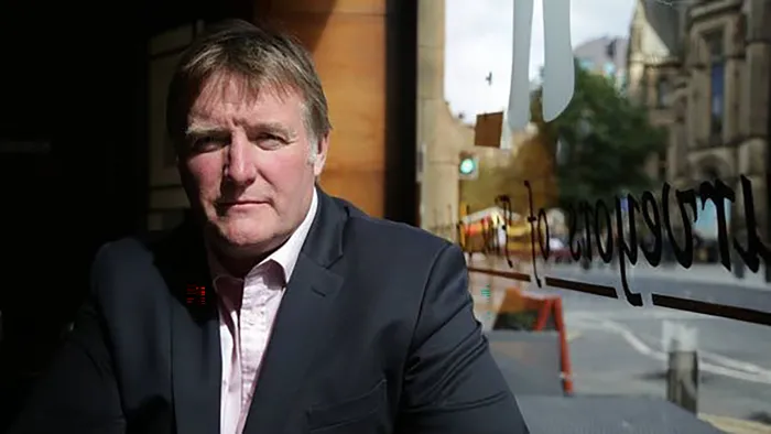 A white man in a black blazer sitting by a window.