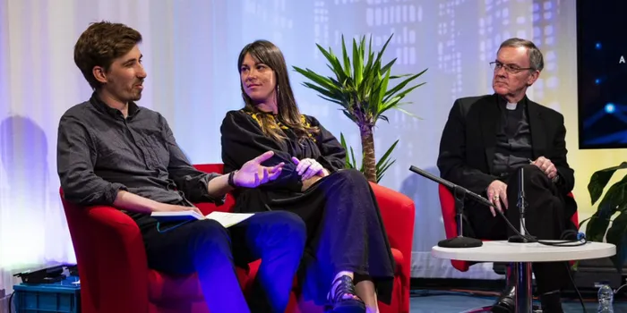 Two men and a woman sit on red chairs talking to each other