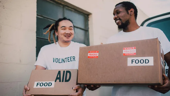 Two men wearing white shirts with the word volunteer on the front and holding boxes of food donations.