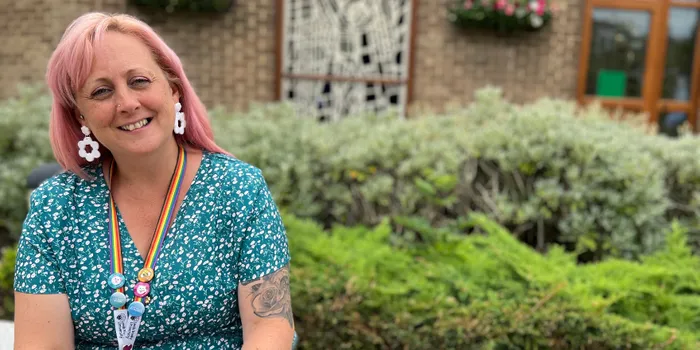 Female with pink hair and green spotty dress sits on bench in front of shrubbery