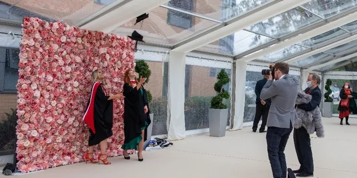 Graduates celebrating in black gowns and caps in front of pink flower wall