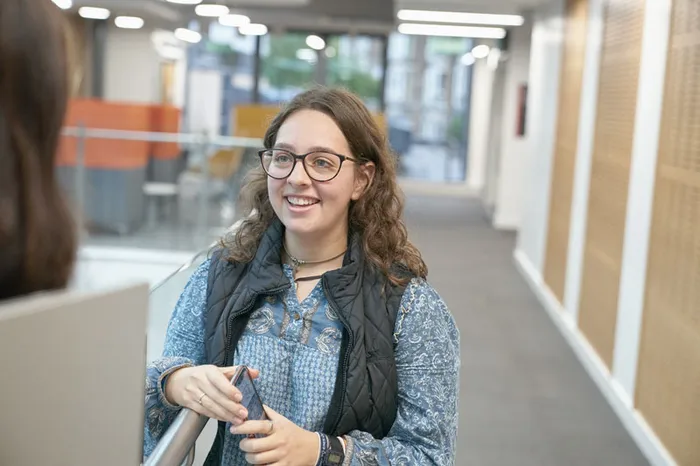 A young woman with shoulder length brown hair and glasses, holding her mobile phone and smiling at someone just off camera.