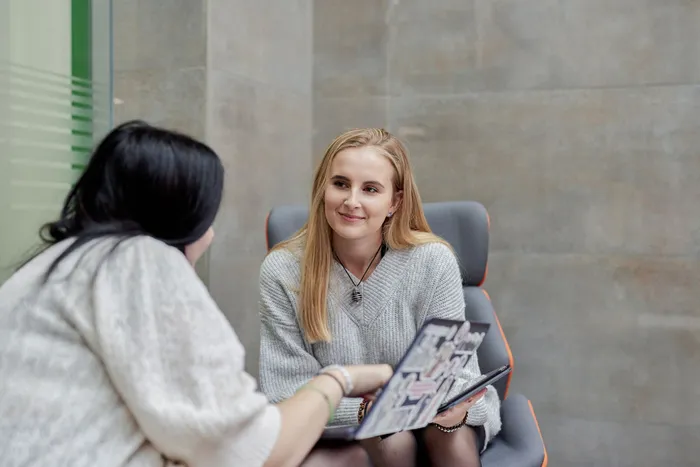 Two students sitting indoors, engaged in conversation; one is holding a laptop covered with stickers while the other smiles attentively.