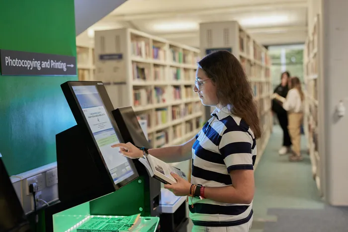 Student in a striped polo shirt using a self-service photocopying and printing machine in a library, holding an open book, with bookshelves and two other people in the background.