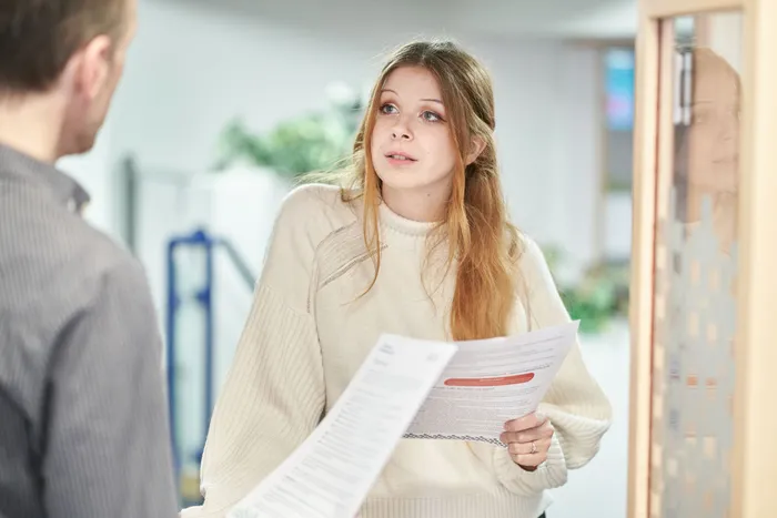Student holding printed information sheets, speaking to another person in an indoor setting, with blurred plants and office furniture in the background.