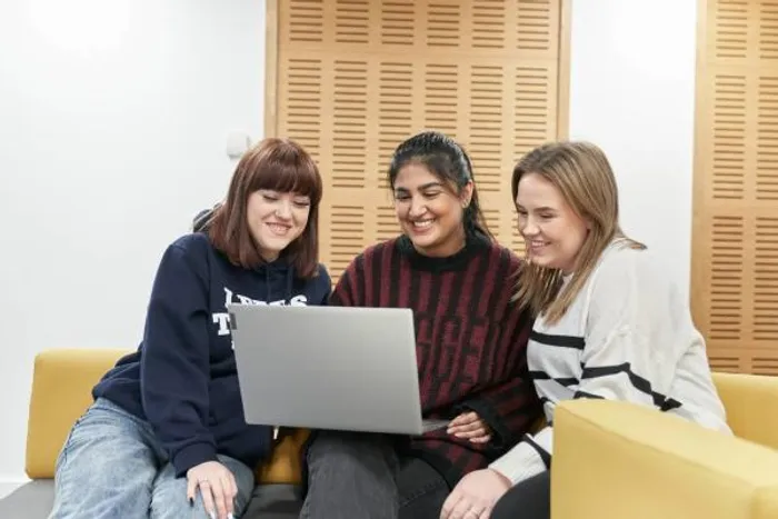 Three people sit together on a sofa looking at a laptop screen in a communal indoor space