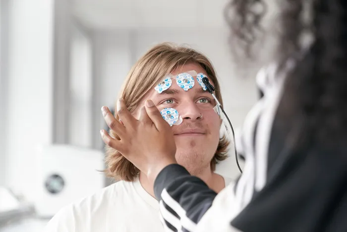 A student with medium-length blond hair sits smiling while another person applies adhesive electrode pads to their face for a psychology experiment. The pads are connected to wires, and the background shows a bright, modern lab space.