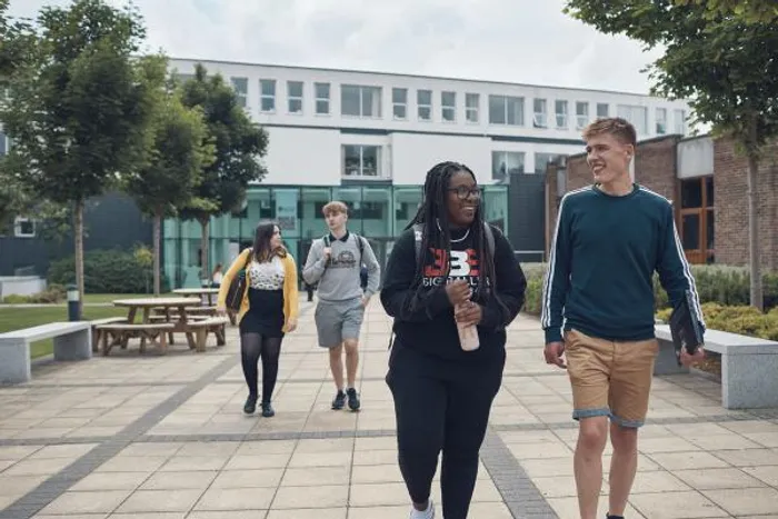 Four university students walk together through a campus courtyard, smiling and chatting. Two students are in the foreground, while two others walk behind them. The area is surrounded by trees, benches, and modern buildings with large windows.