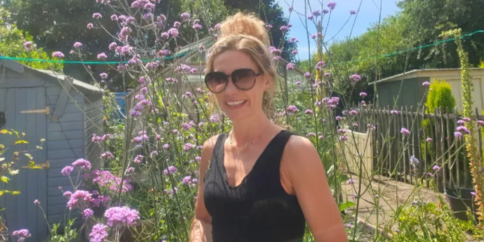 Female wearing black vest top stands in front of flowers with sunglasses on in sunshine