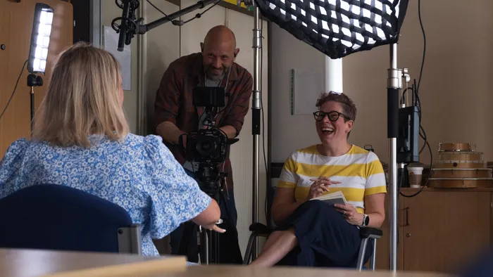 A female academic wearing a striped white and yellow top interviewing another female academic wearing a blue dress for a short film. There is a male camera operator on the left side of the image.