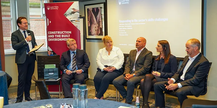 A panel of men and women sitting down in front of a branded Leeds Trinity University banner and having a conversation.