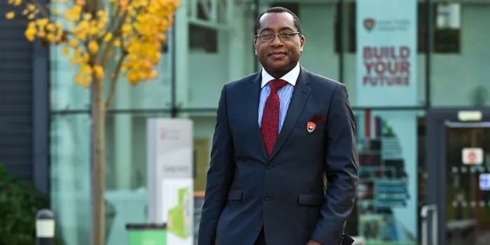 Professor Charles Egbu stands in front of Leeds Trinity University atrium in suit with blue shirt and red tie