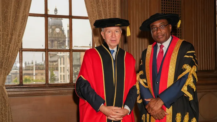 Leeds Trinity University's Chancellor, John Studzinski CBE, on the left, and Vice Chancellor, Professor Charles Egbu, on the right, posing at Leeds Civic Hall in red and black academic robes. Leeds Town Hall can be seen in the window behind them.
