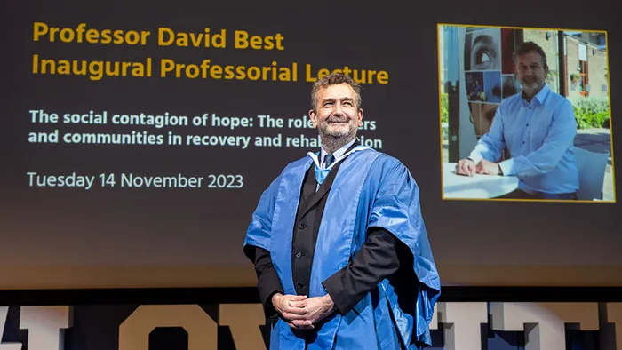 A male academic in a blue gown posing in front of a screen for his Inaugural Professorial Lecture.