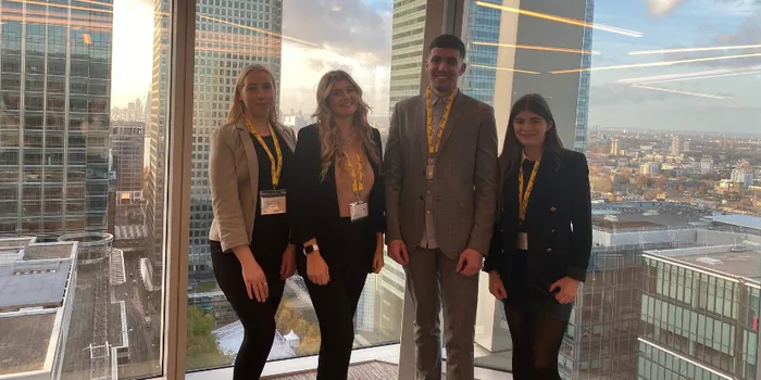 Four students, two blonde females, one brown haired male and brown haired female, stand in front of window showing Canary Whard in London
