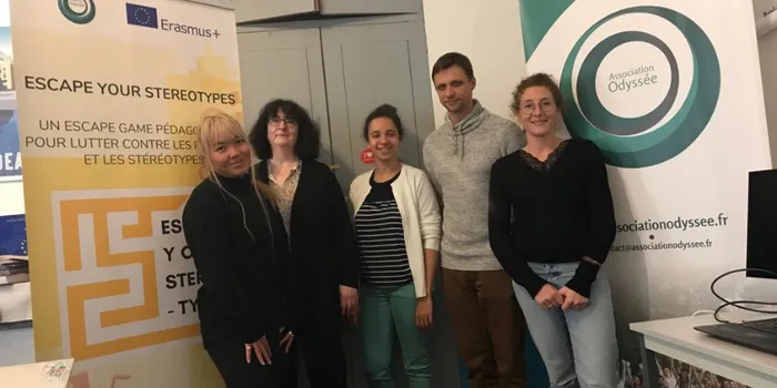 Three women stand to the left of a man in grey jumper with another woman in black jumper to the left in front of two signs