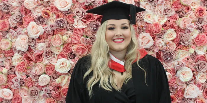Blonde female wearing graduate cap stands in front of flower wall