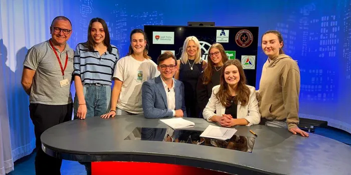 Group of male and female students and tutors stand behind red desk in front of television screen
