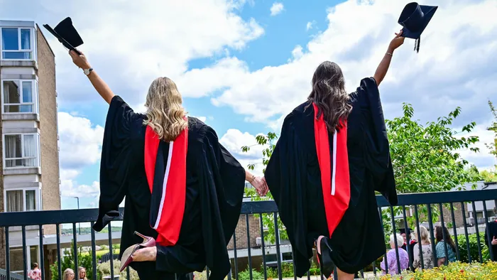 Two students in graduation gowns, holding their caps in the air. They have their backs turned and are holding hands.