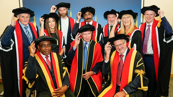 A group of people in Honorary caps and gowns with gold and red accents, posing against a blue background with their hands touching the sides of their caps in greeting.