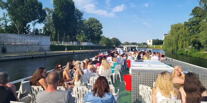 A boat tour in Berlin, with people sitting on a green deck. The river is lined with trees and the sky is blue on a sunny day.