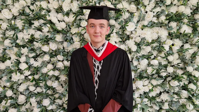 A student in graduation cap and gown smiling in front of a flower wall.