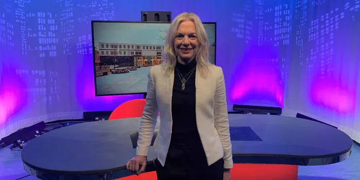 Blonde woman stands in front of broadcast desk and television screen