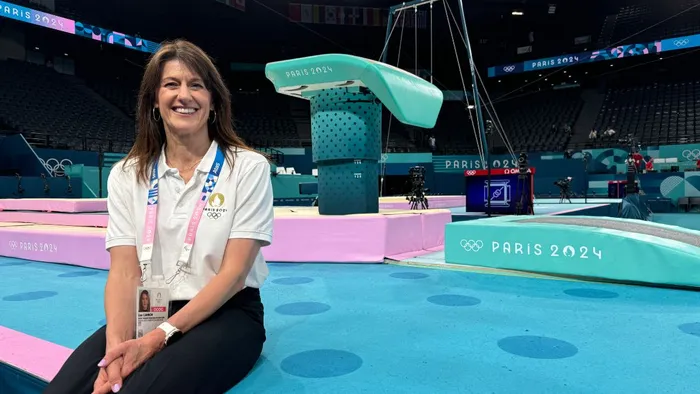 A woman with dark hair and a white top sits on a blue floor in front of gymnastics apparatus