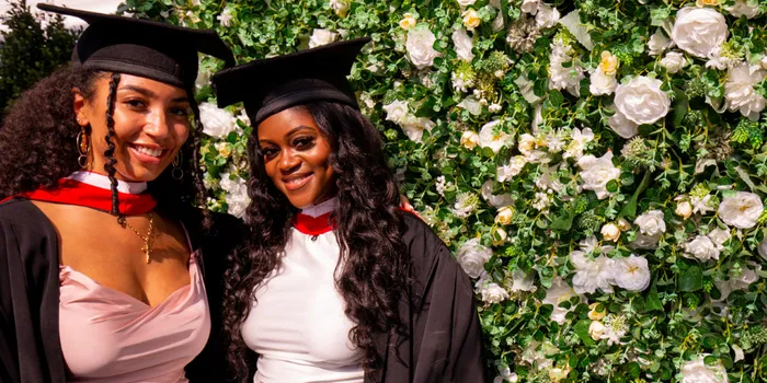 Two female Leeds Trinity graduates stand side by side with graduation caps on in front of a flower wall