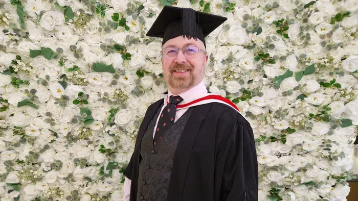A graduate wearing a cap and gown stands in front of a white flower wall