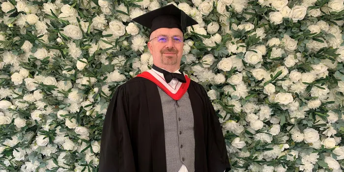 Male graduate stands in front of flower wall wearing graduation cap and gown