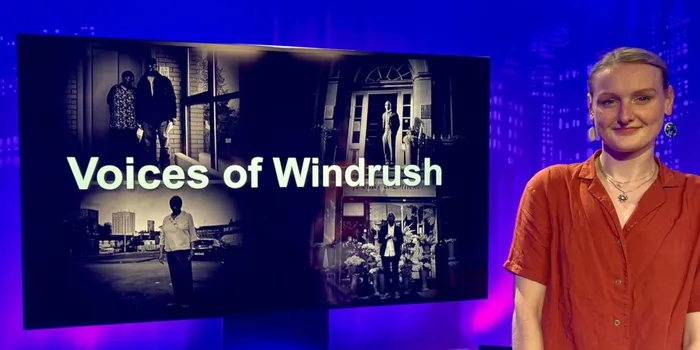 Blonde student in red blouse next to black and white TV screen which reads Voices of Windrush