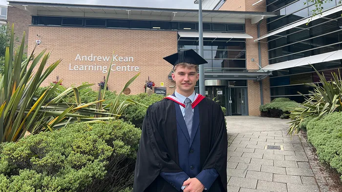 A student in graduation cap and gown smiling in front of the Andrew Kean Learning Centre at Leeds Trinity University.