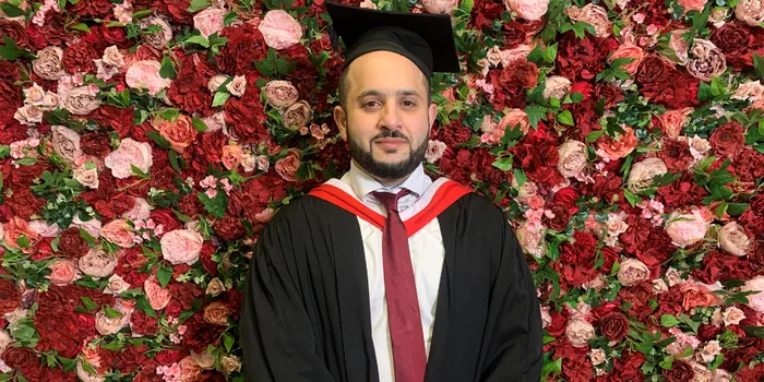 Male in graduation cap and gown stands in front of flower wall