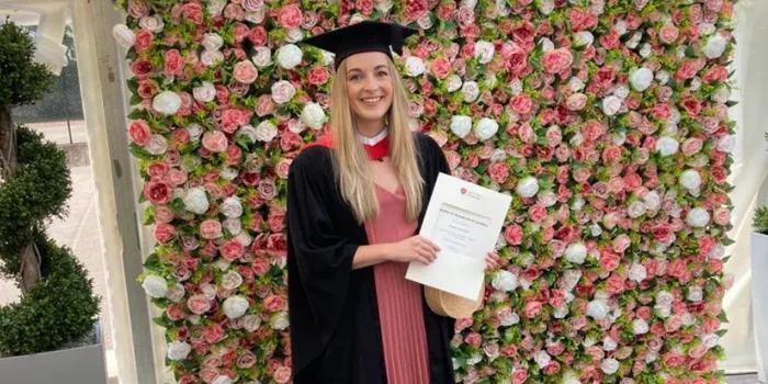 Female student stands in front of flower wall with graduation cap and gown holding certificate