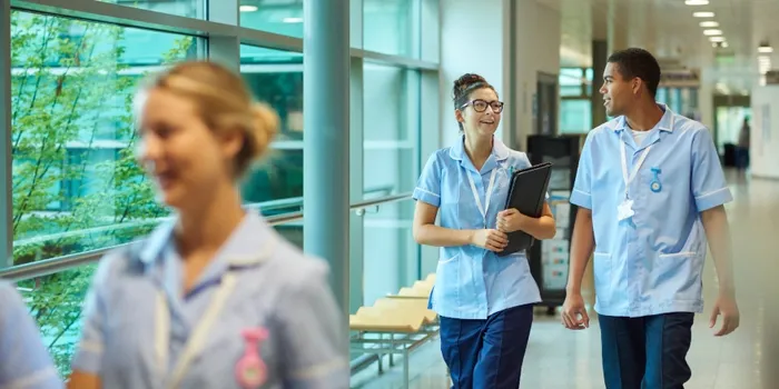 A female and male nurse walking in a corridor, with another female nurse in the foreground.