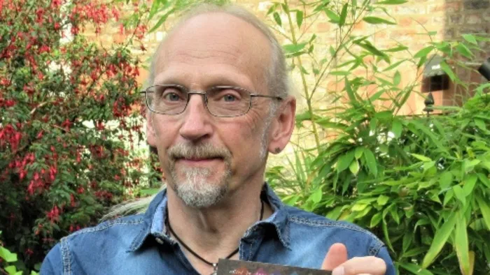 A man wearing glasses and blue shirt smiles while stood in front of green bushes