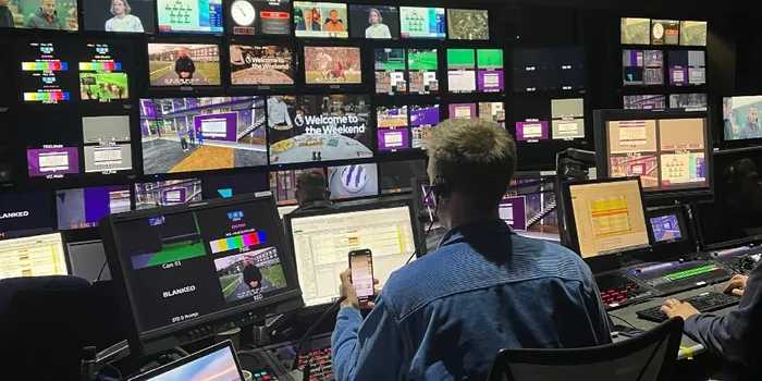 A director in a blue shirt sits at a media production desk, watching a number of different screens
