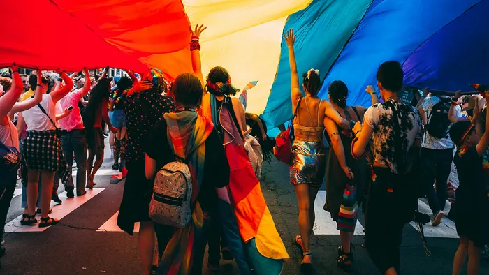 A group of people with their back turned to the camera, underneath a big rainbow flag and holding or wearing rainbow attire.