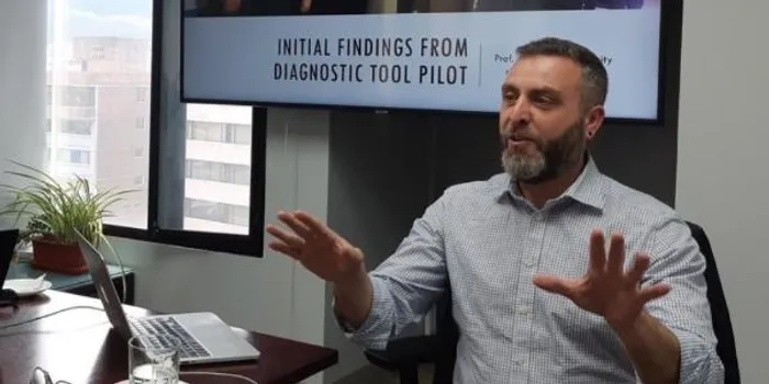 Man with beard sat at a table, actively talking/presenting with a screen behind him