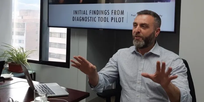 Man with beard sat at a table, actively talking/presenting with a screen behind him