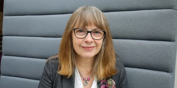 A headshot of a woman with brown hair and glasses, sitting on a grey sofa