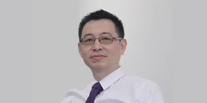 A headshot of a man with brown hair and glasses, sitting in front of a plain white wall