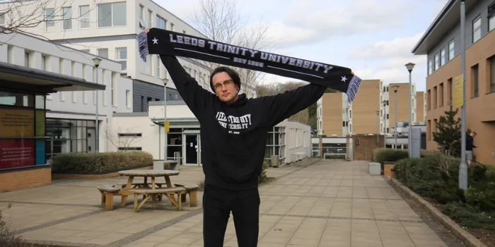 Man with dark hair and glasses stands in front of building holding Leeds Trinity University scarf above his head