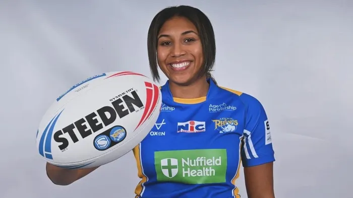 A woman with dark hair wearing a blue top smiles and holds out a rugby ball in front of her