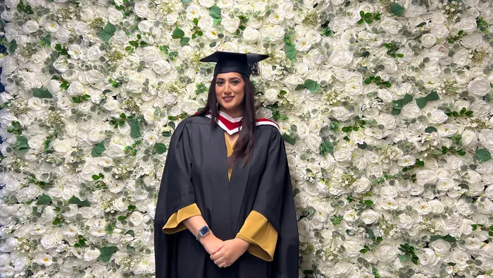 A student in graduation cap and gown smiling against a wall of white flowers.