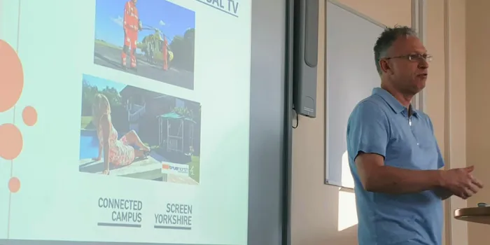 Man with grey hair and sky blue t-shirt stands presenting in front of screen