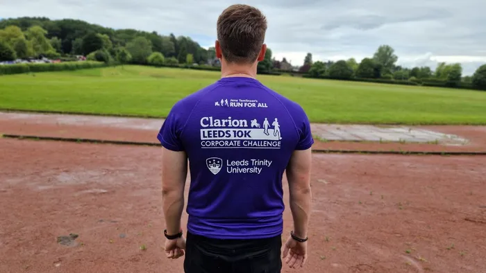 A man with dark hair wearing a purple tee shirt stands on a running track