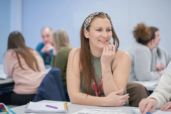 A person with long hair sits at a table in a classroom or study environment, wearing a sleeveless top and a red lanyard. They hold a pen and appear to be thinking or writing. On the table are notebooks, papers, and a purple pen. Other people are seated and working in the background.